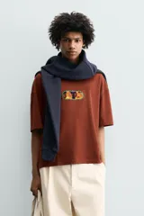 Camiseta de corte relajado color marrón, con cuello redondo y manga corta. Presenta un parche central con diseño gráfico colorido y abstracto.