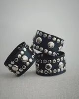 Brazalete ancho de cuero negro con diseño de tachas metálicas plateadas de distintos tamaños dispuestas en filas.