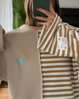 Buzo oversize de corte asimétrico con diseño combinado de rayas verticales y horizontales en tonos beige y blanco. Presenta un corazón bordado en color celeste en el frente y una etiqueta decorativa en la manga.