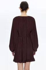 Vestido oversized corto de color marrón oscuro, con escote en V pronunciado y pañuelo haciendo juego que puede abrocharse en la nuca. Cuenta con botones forrados adelante, cinturón fino para atar, mangas globo largas de corte raglán con elástico en los puños y ribete de volados en el bajo.