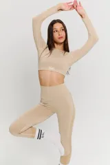 Conjunto deportivo de dos piezas en color beige, compuesto por un top de manga larga con aberturas para pulgares y calzas de tiro alto. Ambas prendas presentan un diseño sin costuras con detalles texturizados y el logo de la marca estampado en el frente del top.