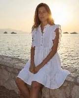 Vestido corto blanco de lino, sin mangas, con volados en los hombros, cuello mao y cierre frontal con botones de nácar. Presenta diseño escalonado con frunce en la falda.