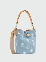 Bolso tipo bucket de denim azul claro con estampado floral blanco. Presenta detalles en cuero color tostado, asa de hombro fija y correa de tela ajustable extraíble. Incluye cierre frontal tipo toggle metálico dorado.