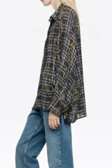Camisa de corte oversize con cuello camisero, cierre frontal de botones y mangas largas con puños abotonados. Presenta un diseño estampado que combina un patrón de cuadros con motivos decorativos en tonos marrones, azules y amarillos.
