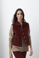 Chaleco de pana color marrón oscuro, con cuello camisero, cierre frontal de botones metálicos dorados y dos bolsillos frontales con detalle de bordado geométrico.