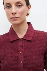 Camisa de manga corta en color bordó, confeccionada en tejido con textura de punto smock. Presenta cuello clásico, cierre frontal con botones y un ajuste entallado al cuerpo.
