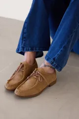 Zapato tipo mocasín acordonado de estilo derby, confeccionado en cuero nobuk color tostado, con costuras visibles y cordones al tono.