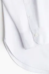 Camisa blanca de algodón con corte holgado, cuello clásico y botones frontales. Presenta mangas largas con puños abotonados y bajo redondeado.