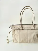 Bolso grande de cuero marrón con textura tipo cocodrilo, estructura rígida y asas para llevar al hombro. Tiene una tira con tachas y hebillas en cada extremo, también lleva lazos en los costados.