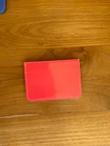 Tarjetero de cuero color coral con acabado brillante, diseño de ranuras en V y placa metálica con el logo de la marca en el frente.