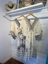 Vestido corto color crudo con bordado floral negro, escote en V, mangas 3/4 acampanadas y cintura ajustable con cordón.