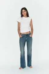 Jeans de tiro bajo con corte bootcut, confeccionados en denim con efecto lavado. Presentan costuras verticales marcadas en el frente, cierre frontal con cremallera, gancho metálico y detalle de trabilla con botones en la cintura.