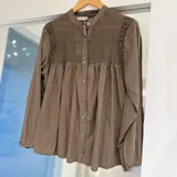 Camisa de manga larga color marrón chocolate, con cuello mao y cierre frontal de botones. Presenta detalles de fruncidos en los hombros y canesú con textura fruncida en el pecho, con diseño suelto y fluido.