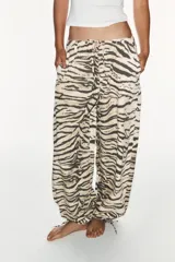 Pantalón de tiro medio con estampado animal print tipo tigre en tonos beige y negro. Presenta trabillas, cintura ajustable con cordones, bolsillos laterales y traseros, y bajo ajustable con cordón. Cierre frontal con cremallera y botón a presión.