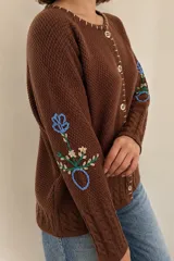 Saco de punto color beige, confeccionado en algodón, con cuello redondo, mangas largas y cierre frontal con botones. Presenta detalles bordados con diseño floral en las mangas y ribetes con rayas azules y blancas en la botonera y el cuello.
