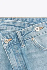 Jeans de tiro alto, color gris oscuro lavado, con pierna ancha extra larga (full length) y diseño de cintura cruzada asimétrica con cierre frontal de cremallera y botones metálicos.