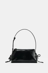 Bolso de hombro de material efecto acharolado negro, con doble compartimento exterior, cierre de cremallera y detalles de lazos en los laterales.