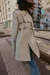 Trench largo de color negro con cuello de solapa, cierre frontal cruzado con botones y cinturón ajustable a tono. Presenta mangas largas con trabillas en los puños, bolsillos delanteros de vivo y abertura en la espalda.