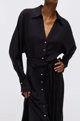 Vestido camisero midi negro de corte holgado, con cuello, escote en V y cierre frontal de botones dorados. Cuenta con mangas largas abullonadas con puños anchos y un lazo ajustable para atar en la cintura.