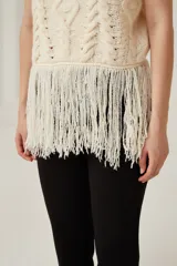 Chaleco de punto sin mangas con cuello polera, diseño de trenzas y rombos en relieve, y detalle de flecos largos en el ruedo.