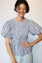 Top de cuello redondo con cuerpo de nido de abeja elástico y mangas globo con elástico en los puños. Presenta un diseño a rayas verticales en tonos azul, blanco y granate, con un bajo estilo peplum.