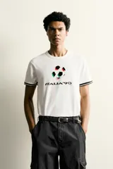 Camiseta de corte relajado confeccionada en piqué de algodón color crudo. Presenta cuello redondo, mangas cortas con ribetes a contraste y estampado frontal con el logo de Italia 90 FIFA World Cup.