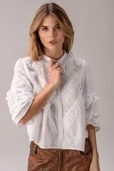 Blusa blanca de broderie con cuello alto con volados, mangas largas abullonadas con detalles de encaje y pliegues decorativos en el frente.