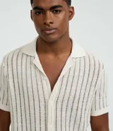 Camisa de manga corta para hombre con cuello solapa y cierre frontal de botones. Presenta un diseño de punto con textura de líneas verticales en color beige y calce regular. Confeccionada en una mezcla de poliéster y algodón sustentable.