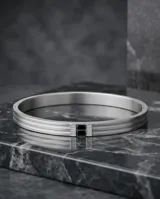 Esclava rígida de acero inoxidable con diseño de tres bandas paralelas y detalle central con piedra de circonia negra flanqueada por pequeñas incrustaciones brillantes.