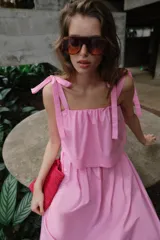 Vestido midi de color rosado con escote recto elástico y tirantes anchos con lazos ajustables. Presenta cintura elástica y bolsillos laterales ocultos en la costura.