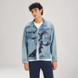 Chaqueta de denim de corte holgado, lavado medio, con bolsillos frontales con solapa, cierre de botones metálicos y detalles de parches e impresiones en tonos más oscuros.