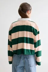 Remera estilo rugby oversize de manga larga y largo cropped, con diseño a rayas horizontales en verde oscuro y rosado. Posee cuello camisero blanco en contraste, cartera de botones, pequeño bordado en forma de corazón en el pecho y terminación desflecada en el ruedo.