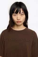 Camiseta de manga corta y escote redondo, color beige claro, con corte boxy. Presenta una hilera de tachuelas metálicas negras decorativas en la parte superior del pecho y en las costuras de los hombros.