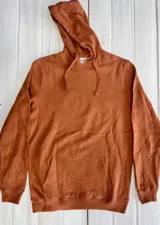 Hoodie color terracota/óxido con capucha y estampado gráfico en la espalda que representa un paisaje montañoso con un sol naciente o poniente y la palabra "MOUNTAIN" en la parte inferior.
