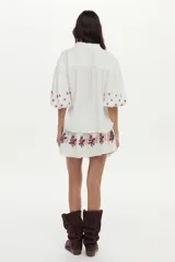 Blusa blanca de gasa de algodón con mangas 3/4 abullonadas y escote en V con botones. Cuenta con bordados en tonos marrones y naranjas en la zona frontal y las mangas.
