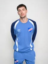 Buzo deportivo de manga larga, color principal rojo, con mangas azul marino y detalles en blanco que bajan desde el hombro hasta el puño. Presenta cuello redondo, logo Umbro y escudo del Club Nacional de Football en el pecho.