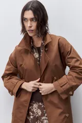 Trench clásico color marrón, de silueta relajada, confeccionado en tejido liviano, con cuello de solapa, doble abotonadura frontal y mangas largas con presillas regulables en los puños.