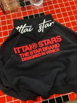 Hoodie negro de corte oversize con capucha, confeccionado en algodón. Presenta un estampado gráfico en color rojo en la espalda con la leyenda 'ITTAI STARS THE STAR BRAND DESIGNED IN VENICE' y un detalle bordado en blanco en la parte posterior de la capucha.