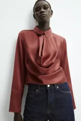 Blusa de tejido satinado con cuello solapa, manga larga y detalle de abertura drapeada en el pecho. Cierre frontal con botón forrado.