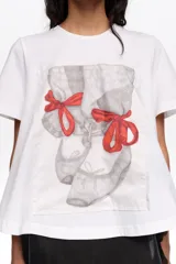 Remera blanca de algodón, de corte línea A con vuelo, escote redondo y mangas cortas. Presenta un estampado frontal con diseño de zapatillas de ballet en tonos grises y lazos rojos.
