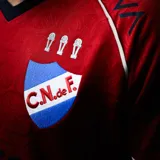 Camiseta de fútbol Umbro del Club Nacional de Football, modelo Away 2026. Diseño en color rojo con patrón de ADN en relieve, cuello en V con detalles en azul marino y blanco, y sponsors estampados en el frente y mangas. Incluye escudo del club bordado con las tres copas intercontinentales.