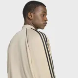 Campera deportiva Adidas Adicolor Classics de corte holgado, color beige con las icónicas tres tiras negras en las mangas y logo del trifolio bordado en el pecho. Cuenta con cierre frontal completo y puños acanalados.
