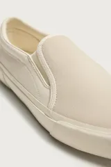Zapatilla casual sin cordones de tipo slip-on, confeccionada en cuero con acabado de serraje y tejido en color beige. Cuenta con elásticos laterales en el empeine y suela de goma a tono.