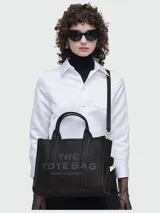 Bolso tote mediano de cuero negro, con asas superiores y correa ajustable y desmontable para llevar al hombro o cruzado. Presenta el logo en relieve "THE TOTE BAG MARC JACOBS" en el frente.