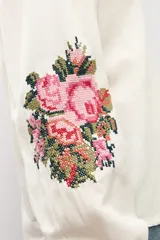 Camisa de color natural con mangas largas abullonadas y bordado floral multicolor en los puños. Presenta cuello mao y cierre frontal con botones.