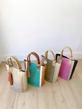 Bolso matero color beige con textura tipo piel de cocodrilo y detalles dorados. Tiene doble asa de mano y un dije decorativo con flecos rosas y marrones.