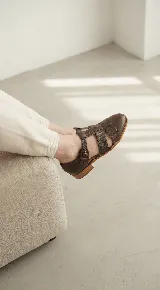 Zapato tipo cangrejera de cuero color marrón oscuro, con diseño de tiras entrelazadas, pulsera con hebilla ajustable al tobillo y taco bajo de madera.