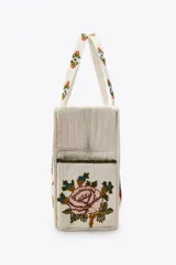Bolso tote acolchado de color blanco crudo, con asas de hombro y bordados florales grandes y coloridos en tonos rojo, marrón, azul y verde. Presenta un forro interior y cierre de cremallera metálica.