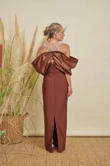 Vestido largo color marrón de corte recto, con escote profundo adornado con pedrería brillante. Presenta un diseño de hombros descubiertos con mangas voluminosas tipo lazo o drapeado, confeccionado en tela satinada.