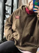 Campera tipo hoodie confeccionada en tela sherpa color verde oliva, con diseño de apliques bordados en la espalda que representan una flor y una fruta. Presenta capucha integrada y corte holgado.